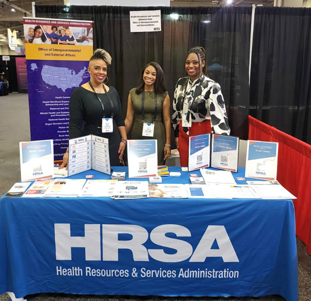 Three women standing behind a HRSA exhibit table at an event.