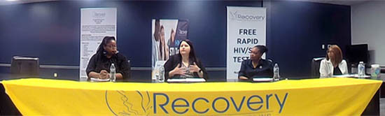 A group of panelists seated at a table covered with a banner stating Recovery.
