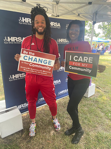 Ryan White HIV/AIDS Program staff hold up signs saying "Be the change in your community" and "I care for my community" at the Baltimore AFRAM Festival.