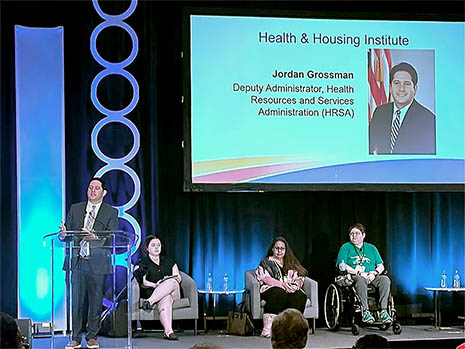 HRSA Deputy Administrator Jordan Grossman speaks at a podium on stage at the Advancing Housing, Health, and Social Care Partnerships Conference.
