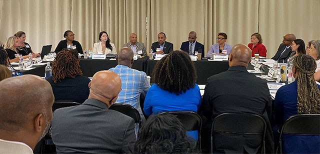 A large group of people is seated around a rectangular shaped table in a formal meeting setting.