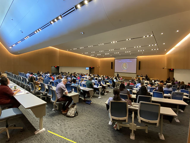 A large group of people is seated in a lecture hall, facing a screen at the front of the room where a presentation is taking place.