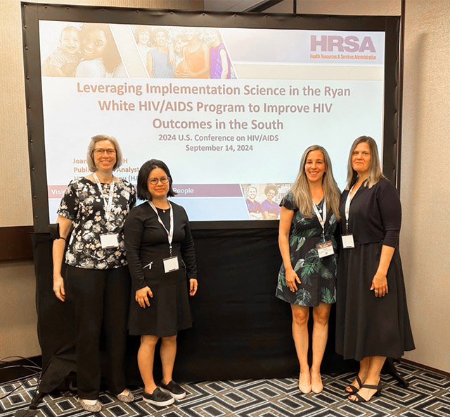 A group photo of four women standing in front of an onscreen HRSA presentation.