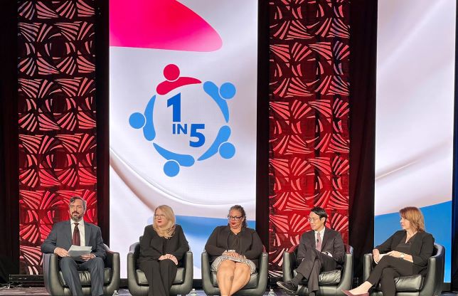 Five panelists, including HRSA Administrator Carole Johnson, are seated on a stage with red and black patterned banners.