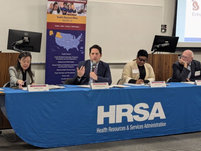 Panel discussion with four HRSA leader seated behind a table covered with a blue tablecloth featuring the HRSA logo.  