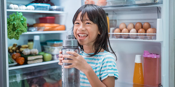 Child standing in front of an open refrigerator holding bottled water.